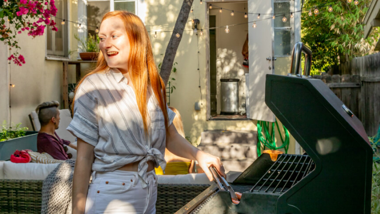 Eine weiße Frau mit roten Haaren und einem weißen Outfit grillt Hotdogs auf einer Party im Freien.