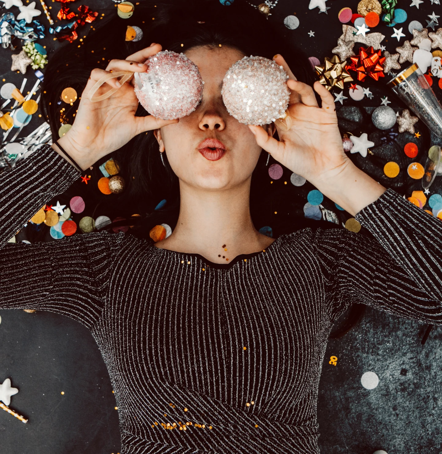 Image of a woman laying on the ground, with two small disco balls covering her eyes and her lips pursed. Confetti covers the ground around her, and she's wearing a long-sleeved sparkly shirt. 