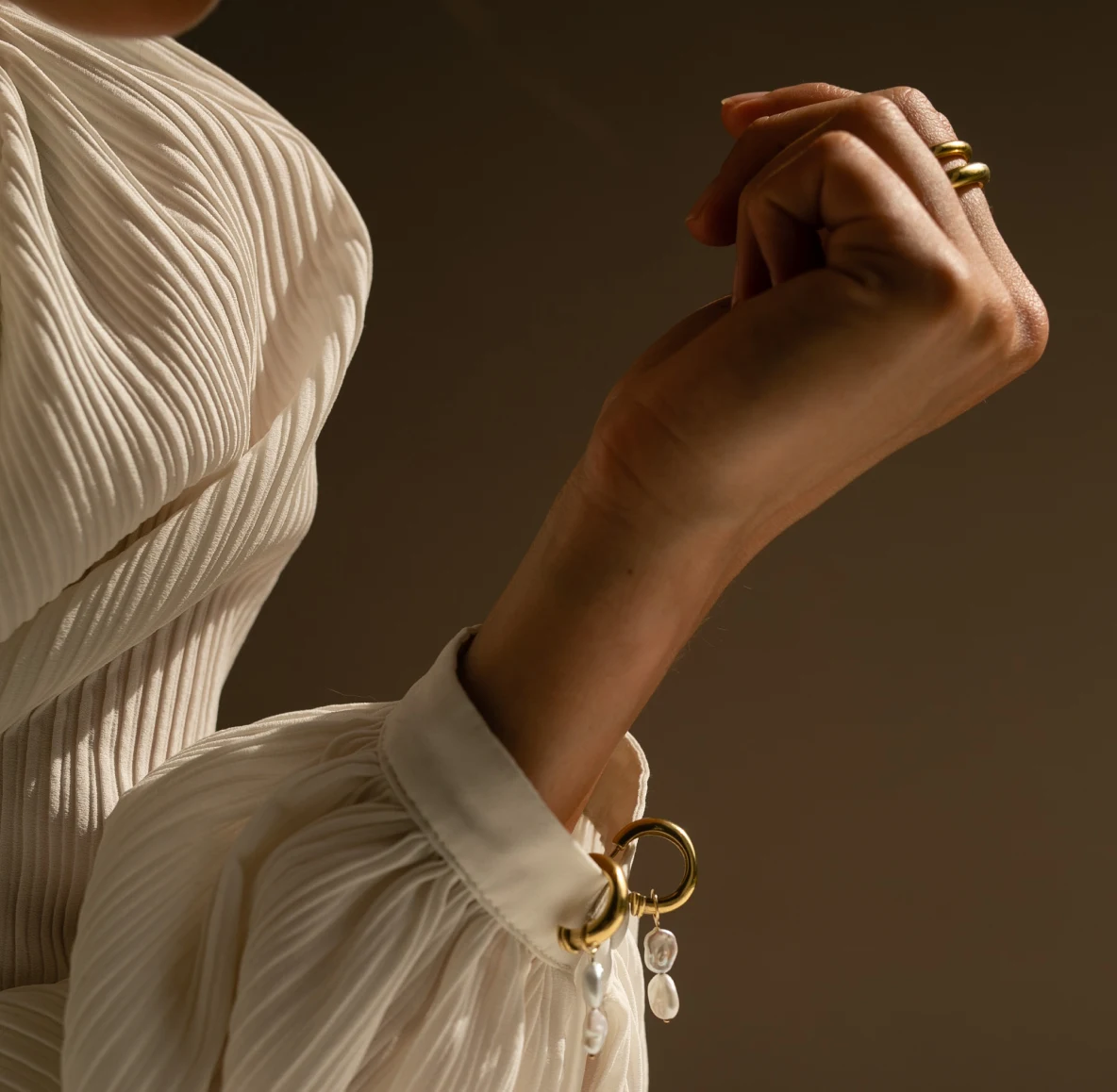 A woman’s arm wearing a pleated white blouse with gold and pearl cufflinks and a thick gold ring on her pointer finger