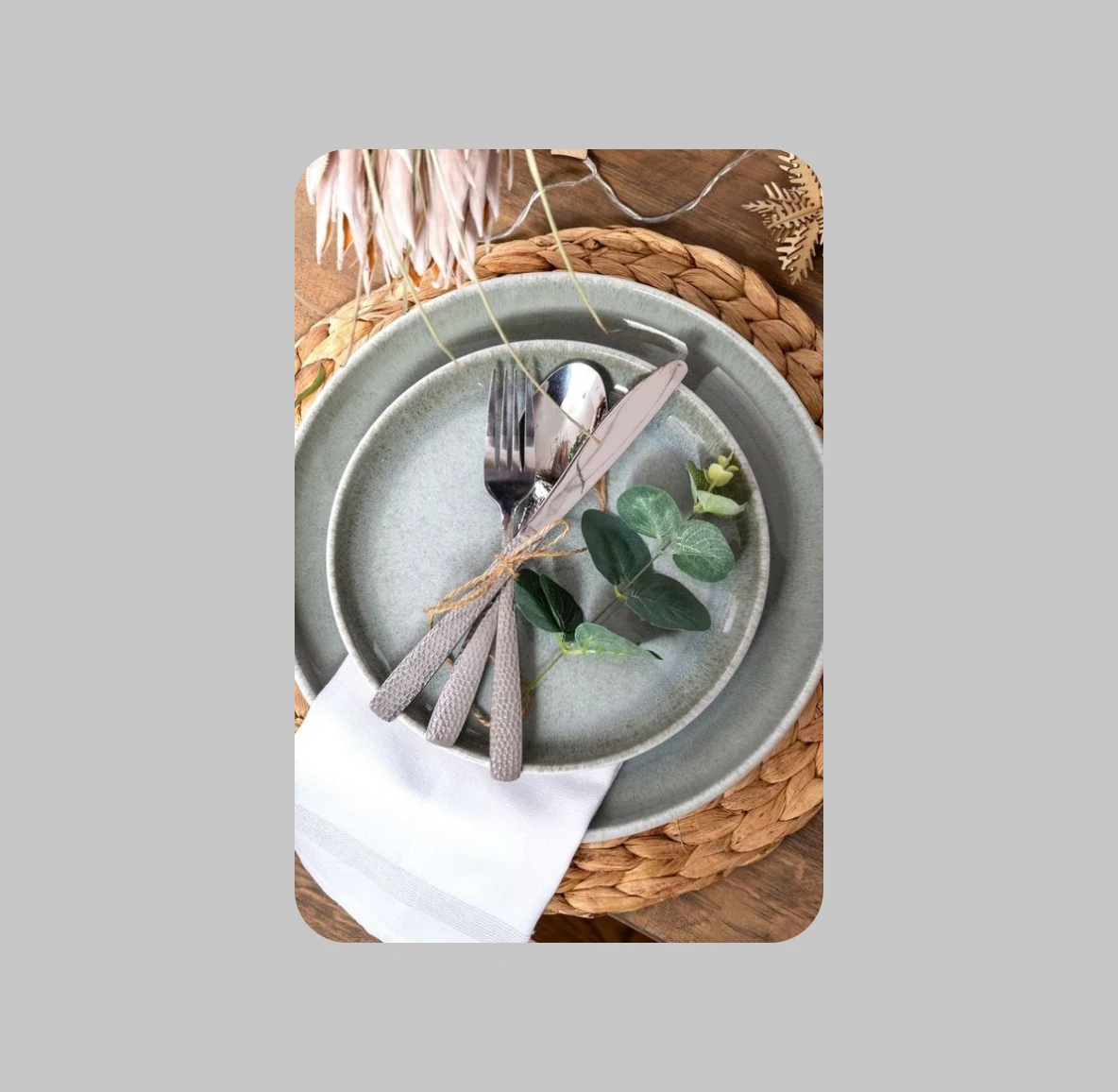 A set of silverware—including a spoon, knife and fork—wrapped in twine and a sprig of eucalyptus is placed on top of two, matching light green plates. Beneath the plates is a woven, wooden circular placemat and a white paper napkin. 