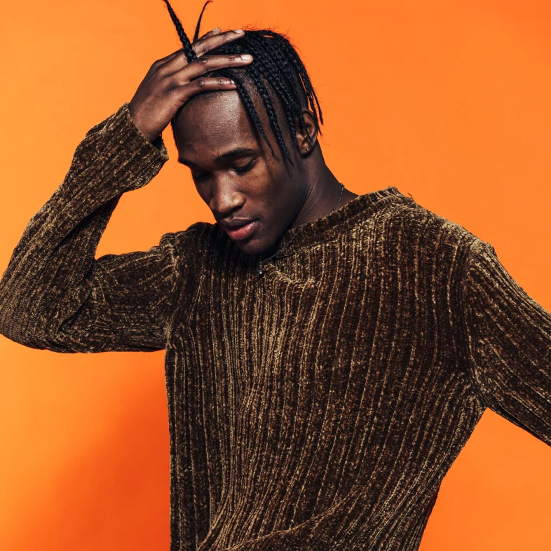 Black man with one hand on his head, holding his hair back out of his eyes. He is clothed in a brown sweater with gold accents. 