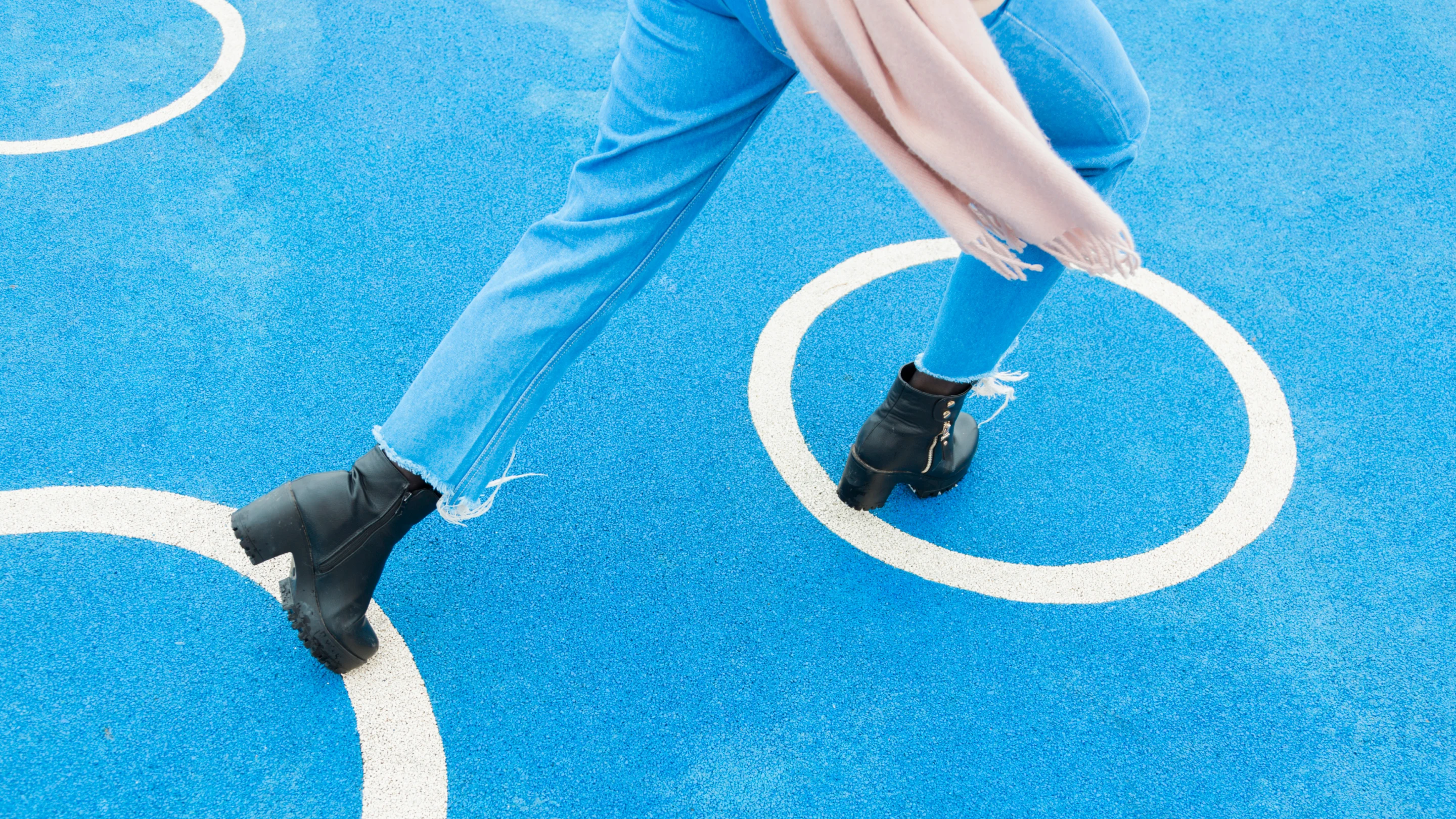 Woman in jeans and black boots runs across blue rubber ground of a playground, over three large white circle, her pink scarf blowing into frame.