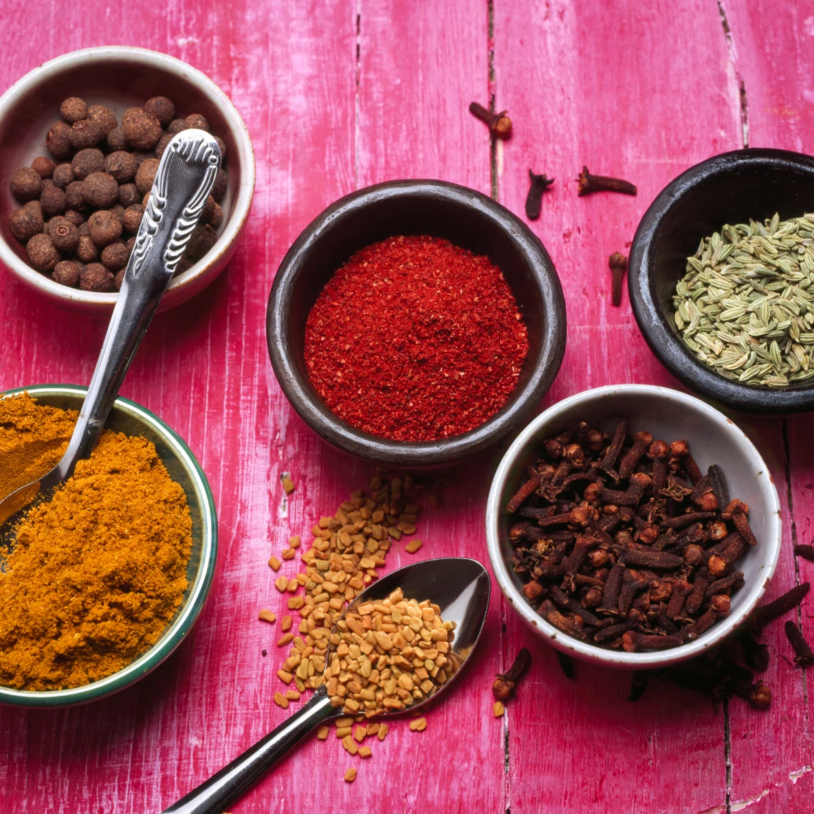 Five bowls of colorful spices and silver spoons placed on a pink wooden table