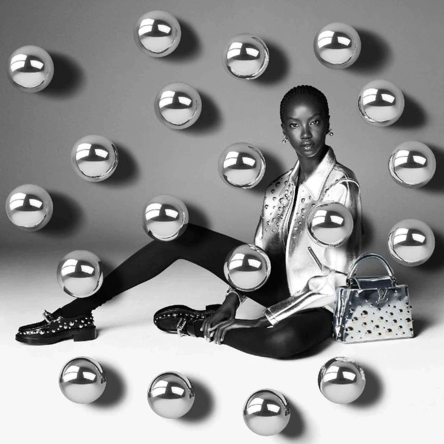 Black and white photo of model styled with Louis Vuitton silver jacket, handbag, and loafers surrounded by hanging silver orbs