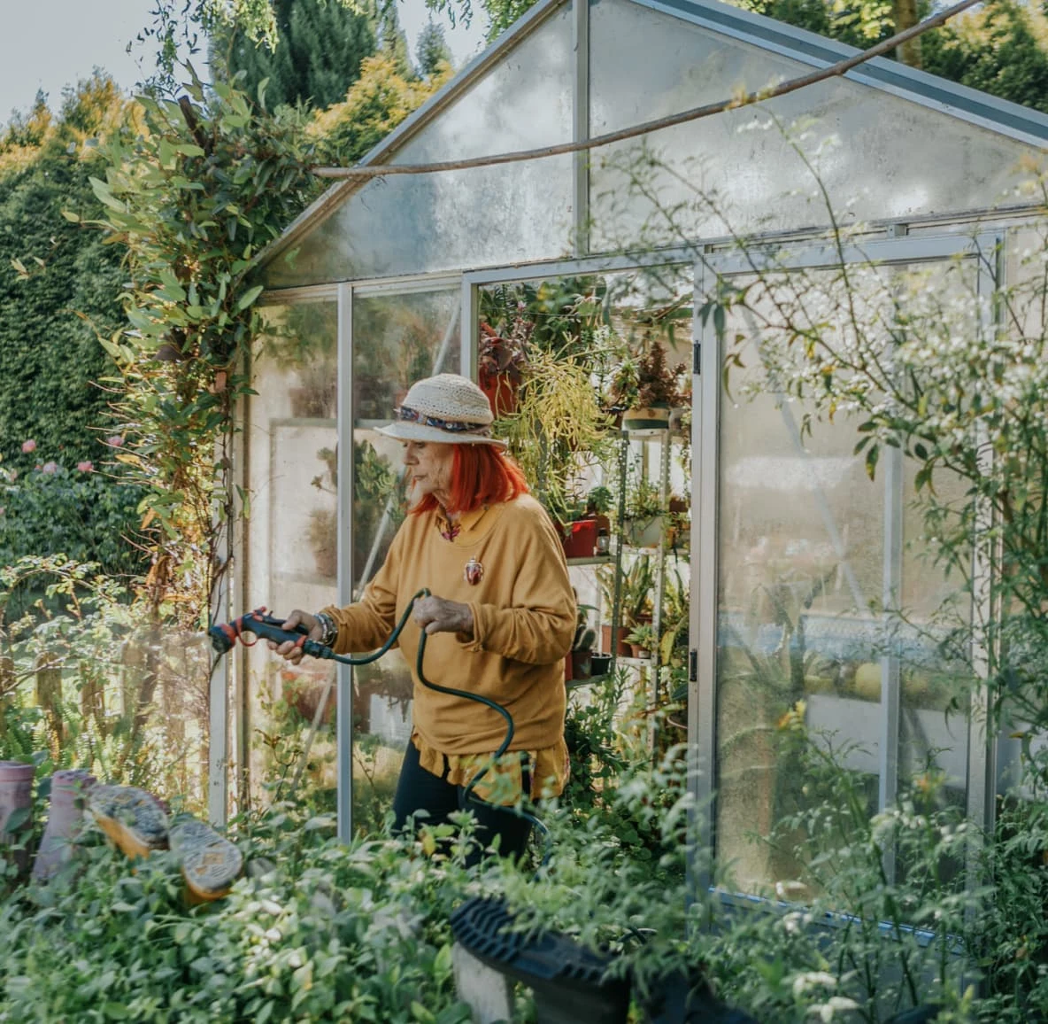 a White woman in a straw hat waters plants outside a greenhouse