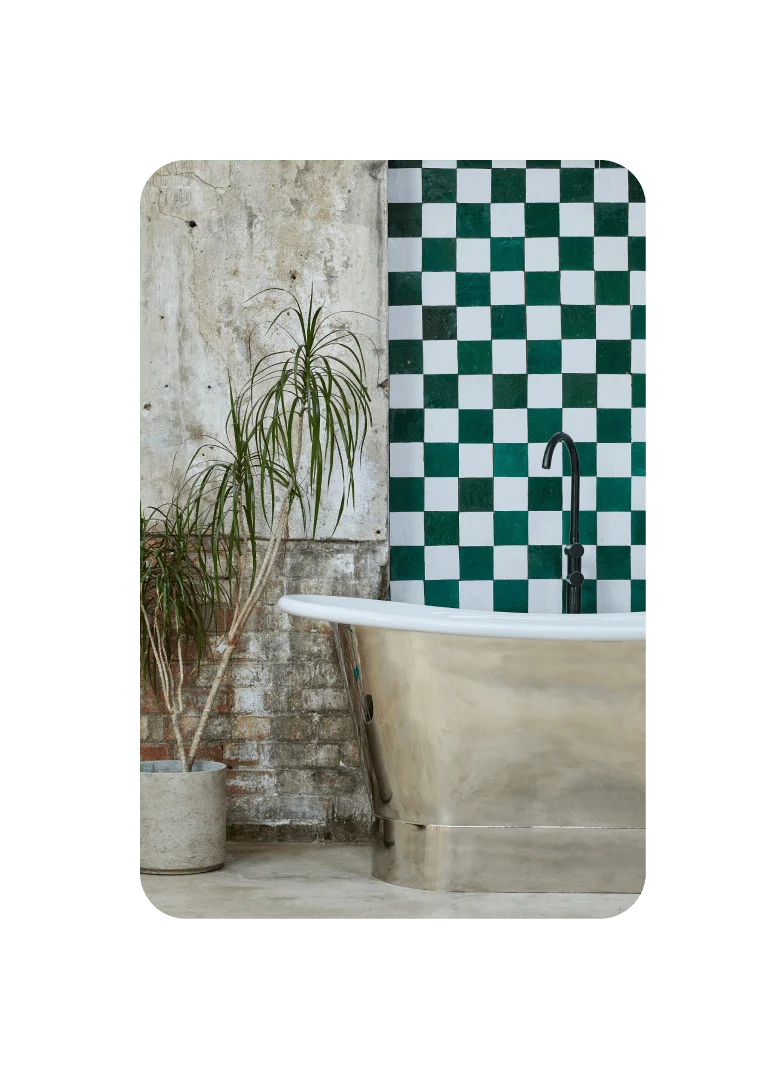 Pin featuring a focused view of a bathroom decorated with a palm plant in a cement pot and metallic tub against a checkered wall.