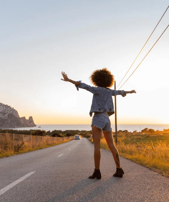 A woman with curly hair, wearing a denim jacket and denim shorts, is standing with her arms out on a country road.