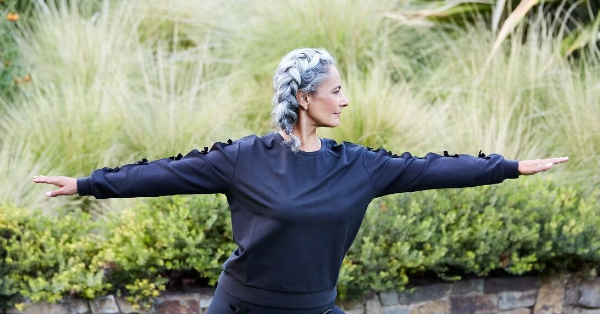 Woman with silver french braid wearing a navy long sleeve top doing warrior pose in front of a lush green background