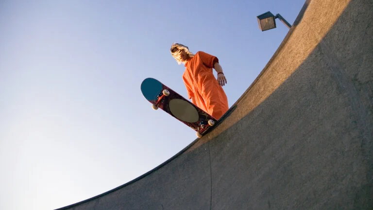 A White man with long blonde hair preparing to descend down a skateboard ramp on a pink and blue skateboard. 