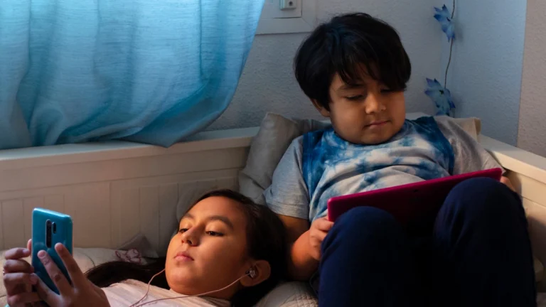 Duas crianças asiáticas deitadas em uma cama, usando dispositivos eletrônicos para se entreter.