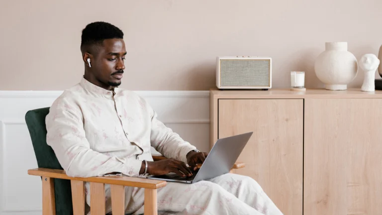 Homem negro vestindo camisa e calça bege e usando fones de ouvido, sentado em uma cadeira e digitando no computador.