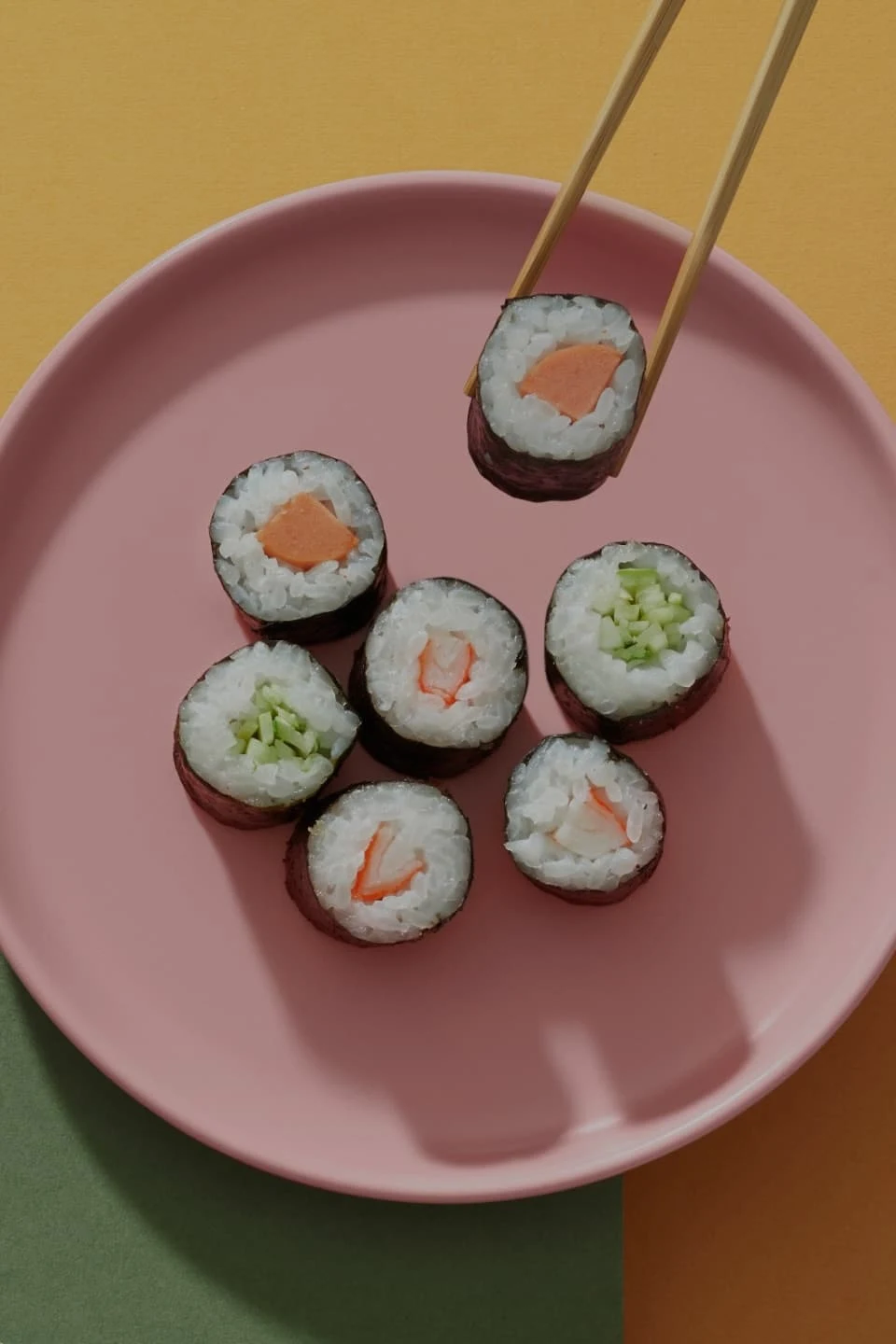 Assorted sushi rolls being grabbed by a pair of chopsticks on a pink plate in front of an orange and green backdrop