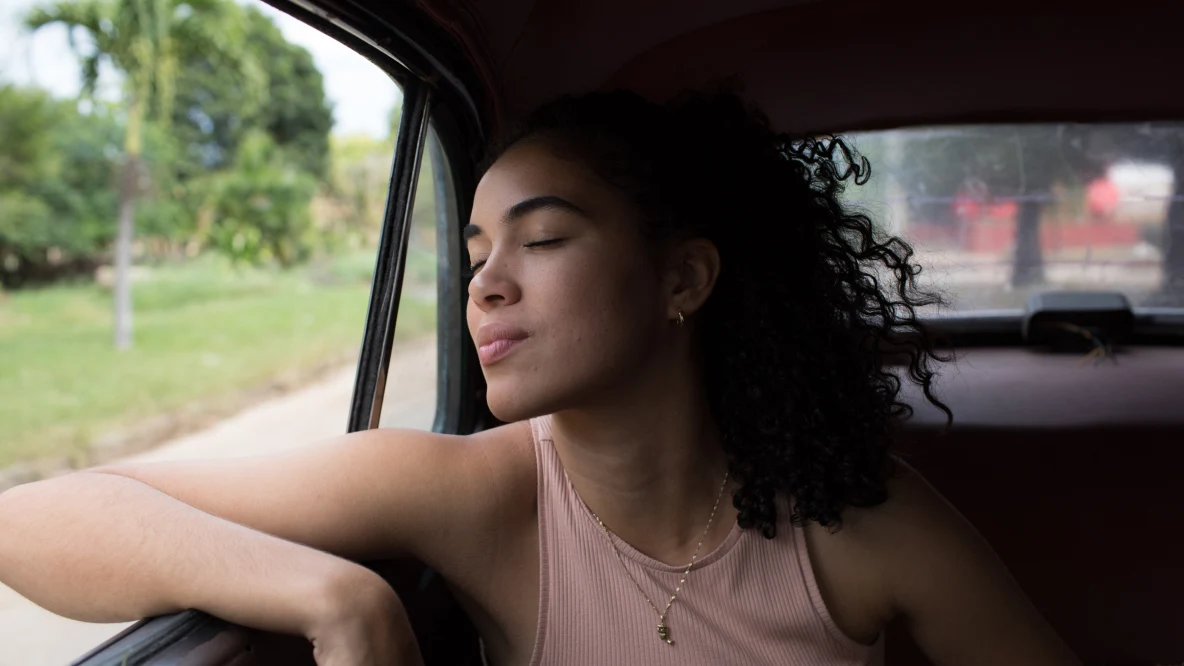 A fair-complexioned Black woman with long curly, dark hair leans an elbow out of a car window, her eyes closed, green grass and trees in the background