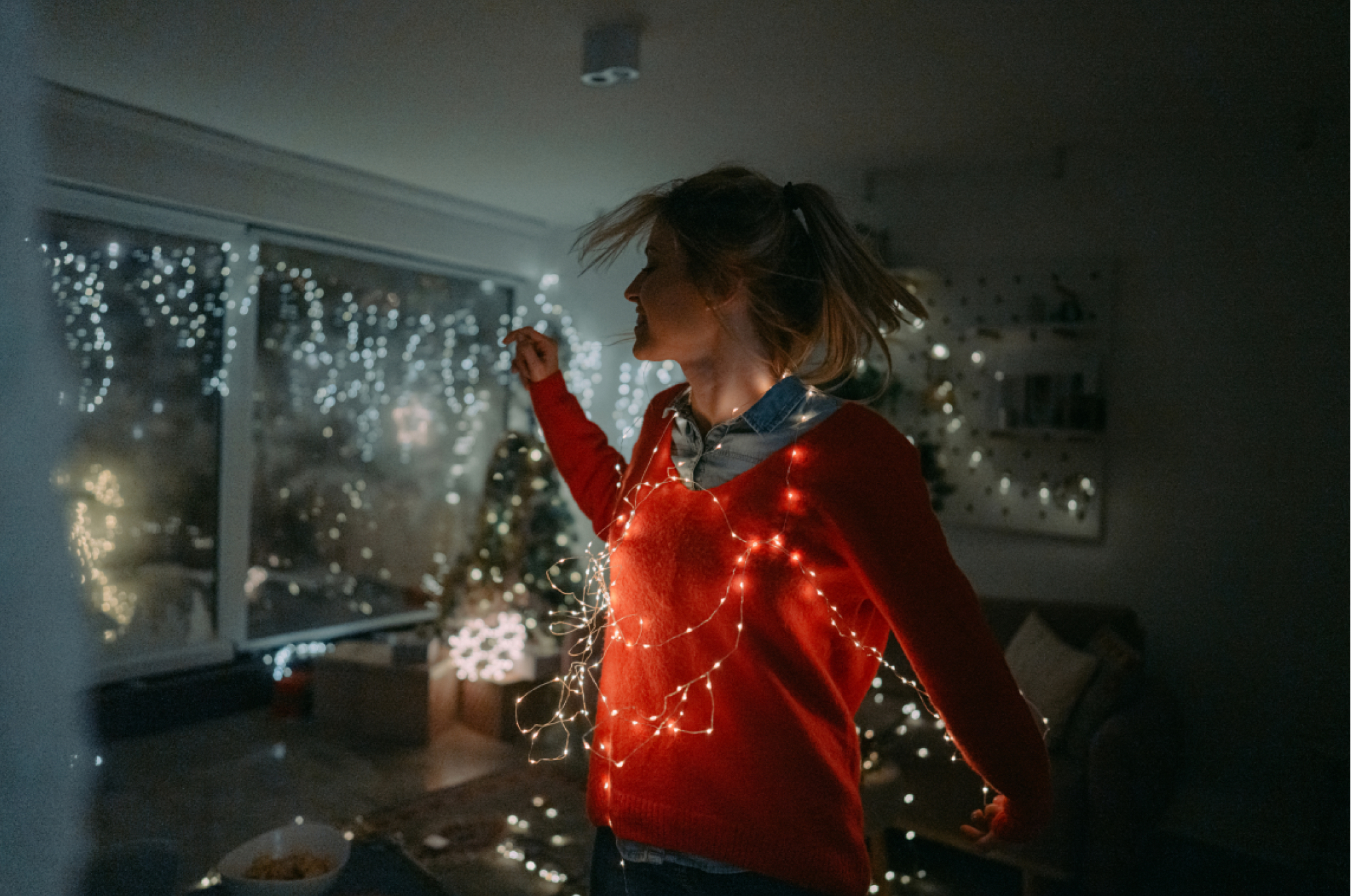 Christmas joy. Photo of a young woman wrapped in Christmas lights and dancing in her well-decorated living room; enjoying and celebrating Christmas eve alone in her apartment.