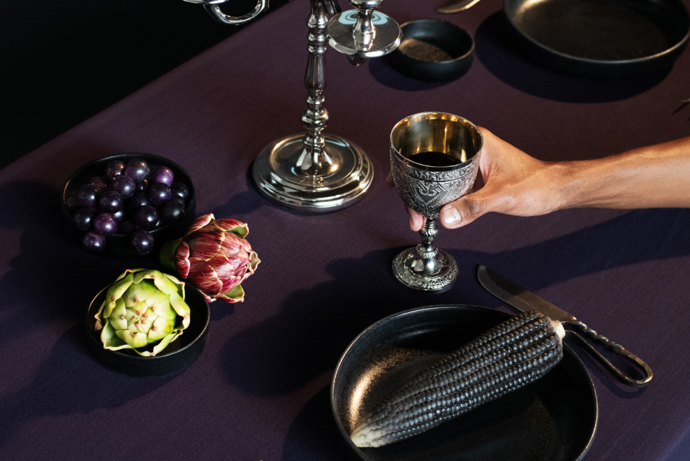 Adark-toned dinner table with silver goblet, artichoke, and grapes, evoking Day of the Dead and Halloween themes