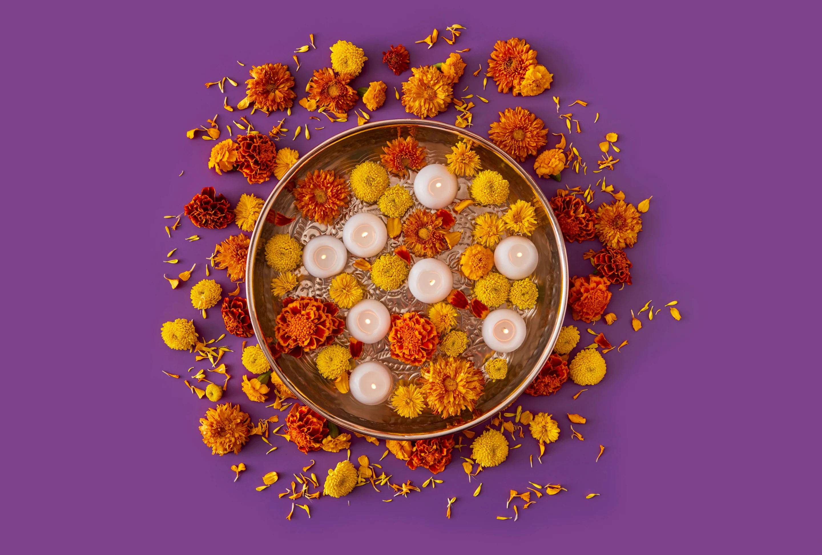 Orange and yellow flowers and white candles floating in water inside a round metal bowl. Orange and red flowers surround it against a purple background.