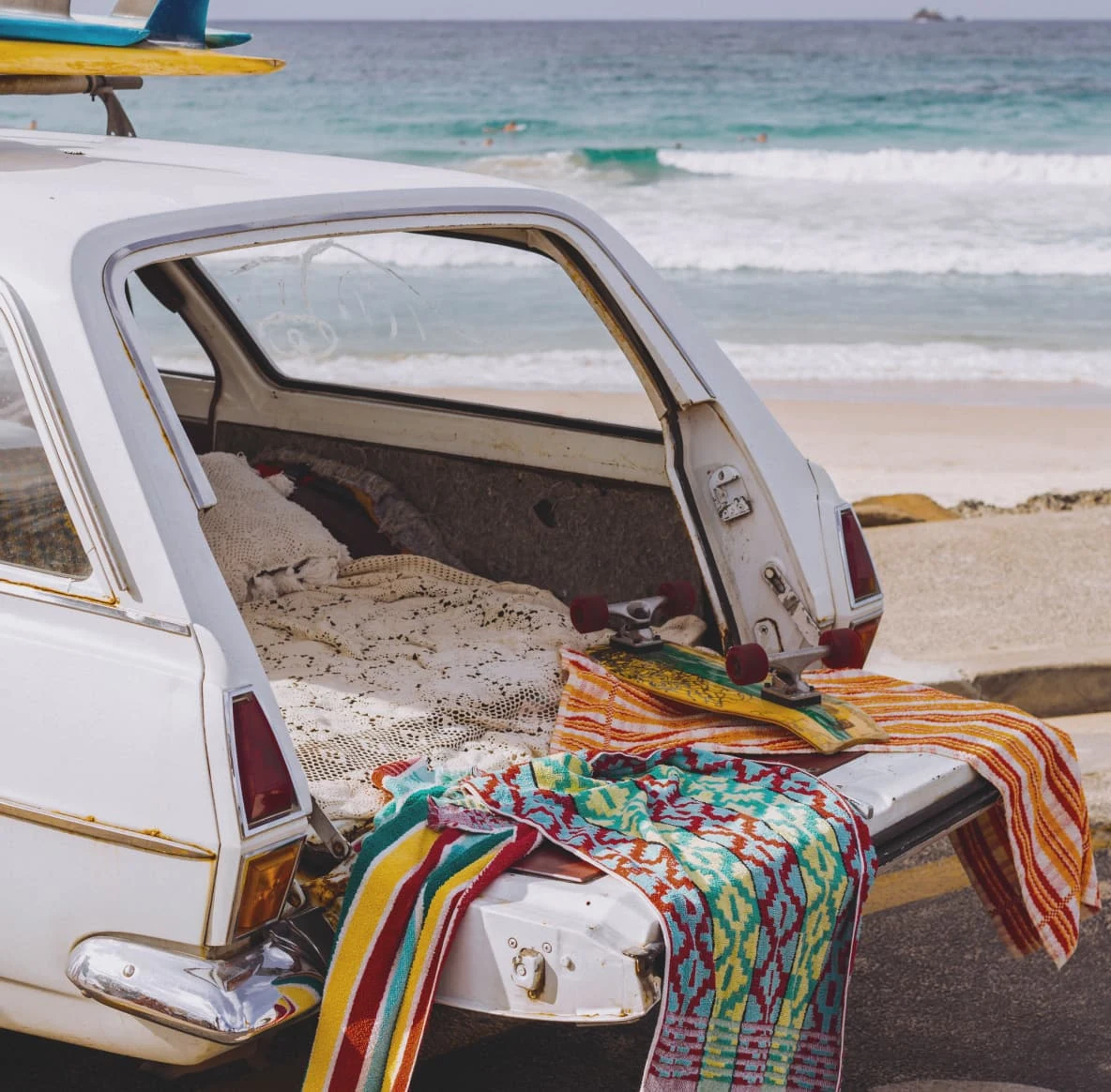 The open back of a white station wagon covered in beach towels, surf boards on top and the ocean in the background