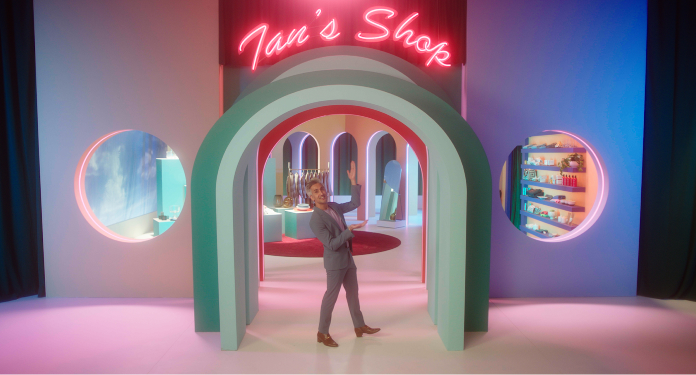 Tan France standing at the light green entrance of ‘Tan's Shop’. The shop's title is written in neon pink. Two circular windows appear to the left and right, giving viewers a glimpse of the shows and clothing items inside. 