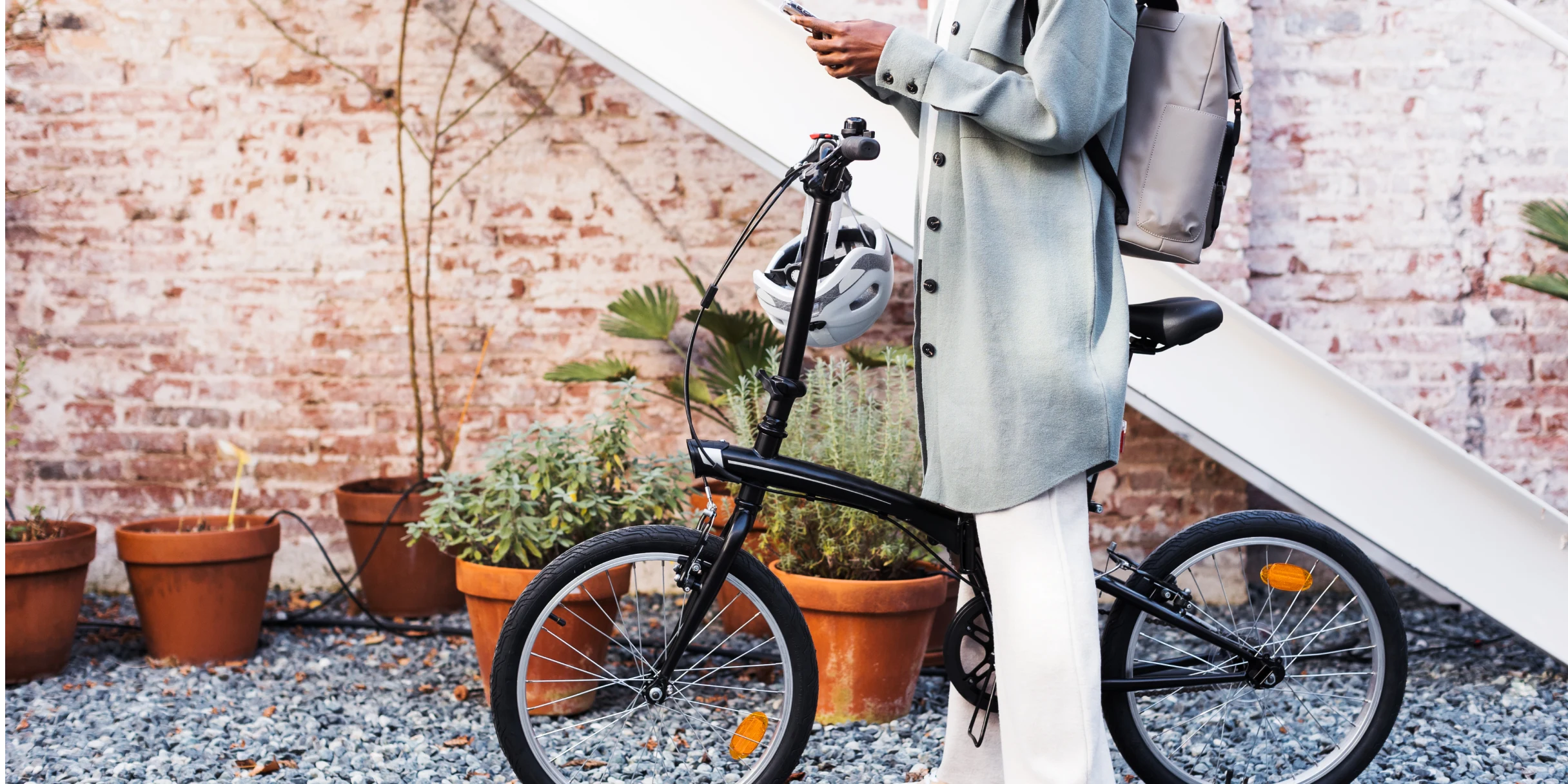 A person in a gray coat stands near a brick wall and straddles a black bicycle