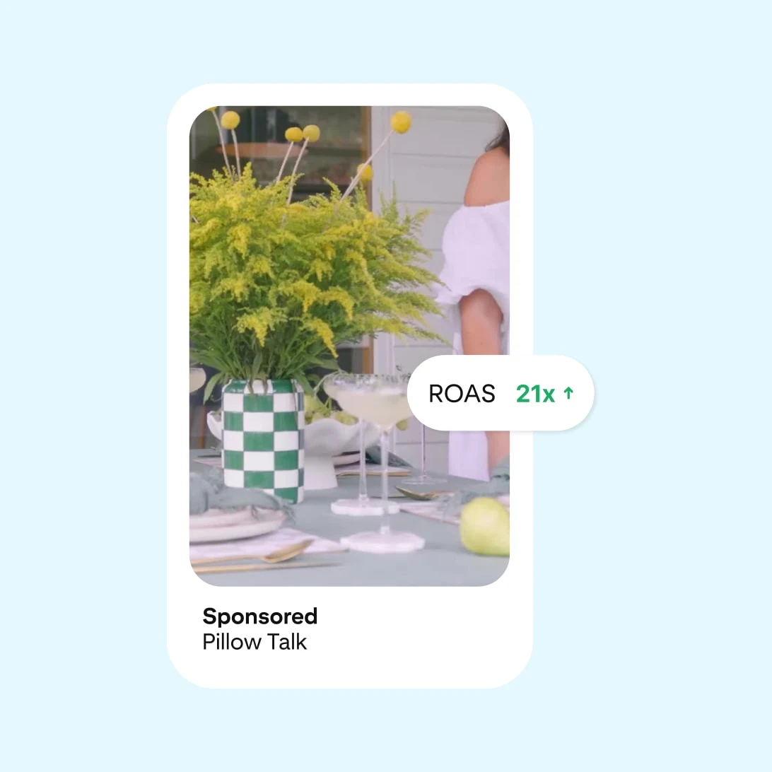 Pinterest ad showing an outdoor dining table set with greenery, Checkerboard details, and glasses. Partial view of a woman in white dress. Includes "ROAS 21x" tag, sponsored by Pillow Talk.