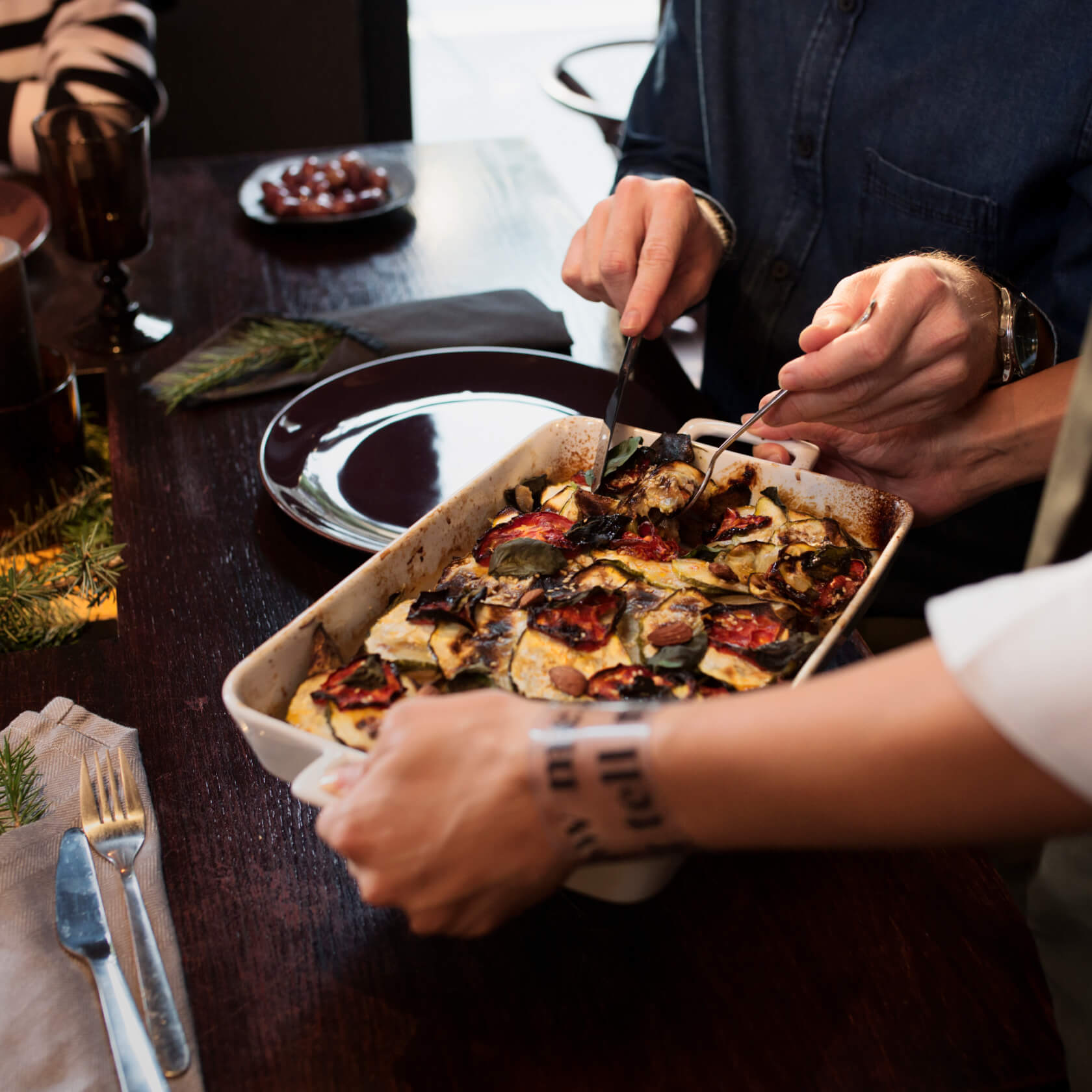 Someone serving dish of roasted veggies at decorated table while second person cuts out a serving with knife and fork