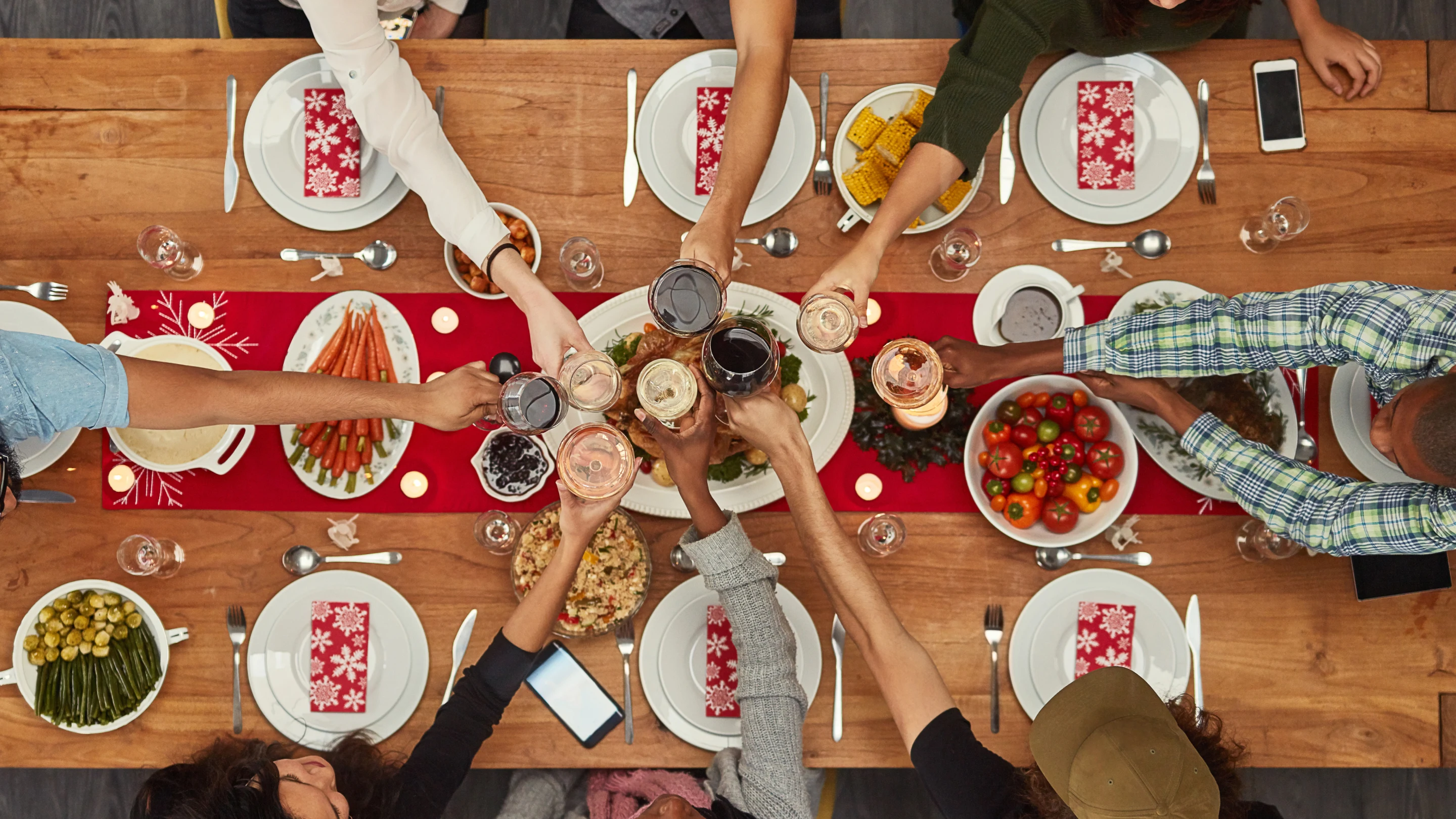 Imagen de una mesa con personas a punto de compartir una comida. La mesa es de madera y tiene un mantel fino de color rojo intenso que se extiende por el centro. Hay siete personas sentadas a la mesa y todas ellas sostienen una bebida para brindar. La mesa está cubierta con una variedad de platos, que incluyen tomates frescos, trozos de mazorcas de maíz, zanahorias, un pollo asado y mucho más. Delante de cada persona hay un juego de platos de porcelana y cubiertos (un tenedor, un cuchillo y una cuchara), y una servilleta floral blanca y roja. 