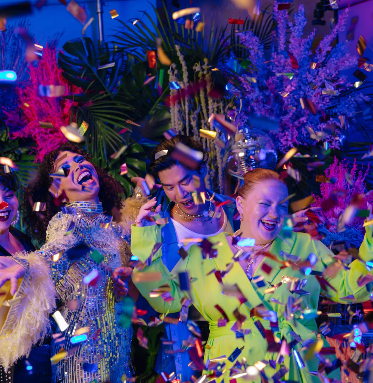 A group of young people dressed in colorful and sparkly party wear, smiling and dancing as confetti falls around them.