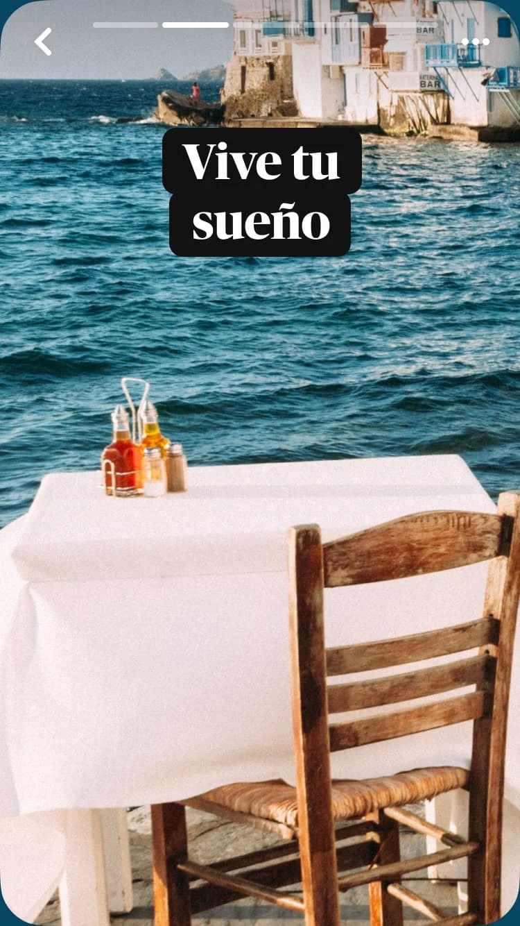Una mesa dispuesta para uno en un café al aire libre, cubierta con un mantel blanco; en el fondo, se ven el mar y edificios, y el texto superpuesto es &quot;Vive tu sueño&quot;