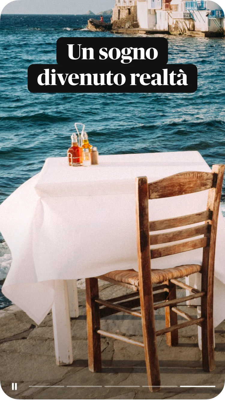 Un tavolo di un bar all'aperto con una tovaglia bianca e una sola sedia rivolta verso il mare e dei tipici edifici greci, con una sovrapposizione di testo che dice "Un sogno divenuto realtà"