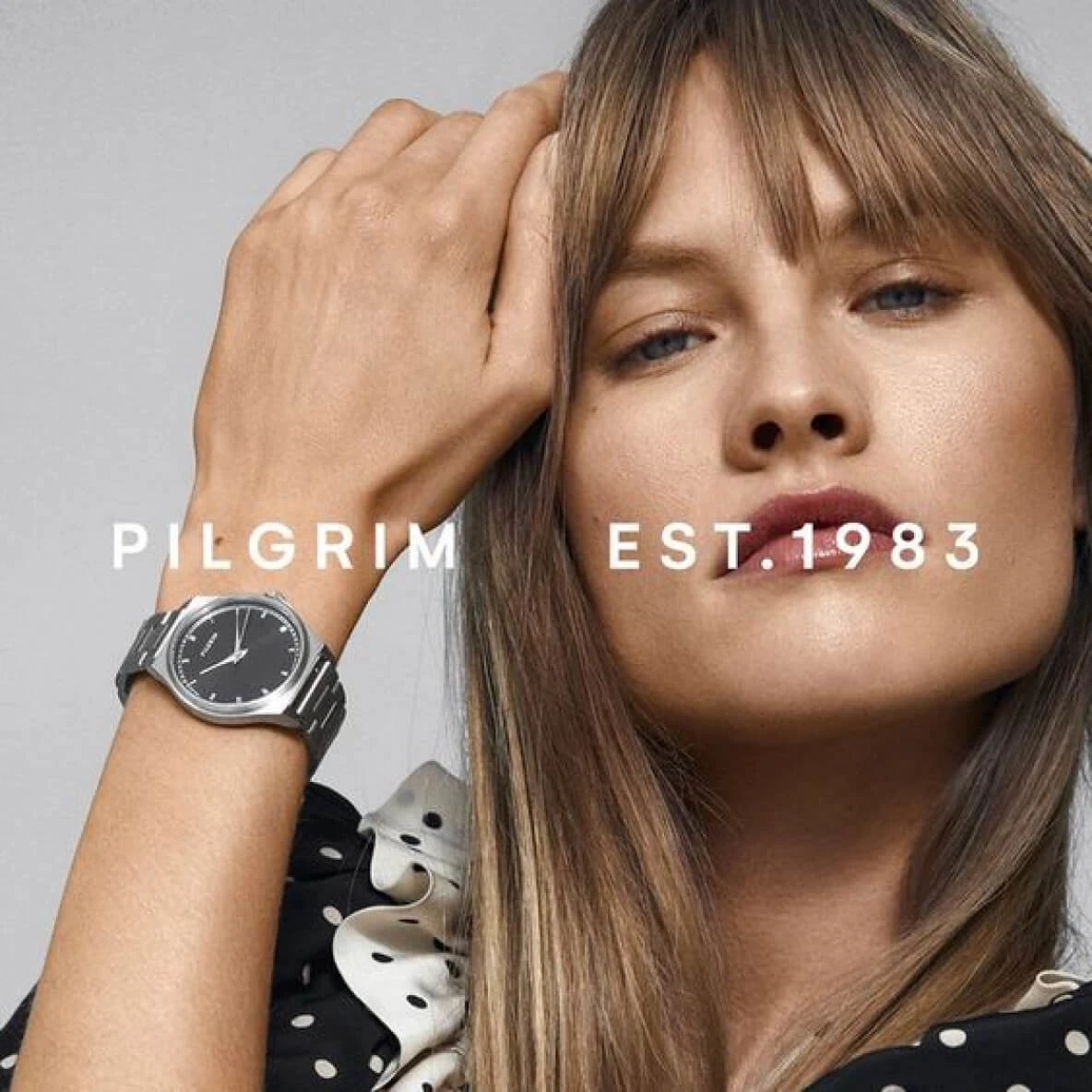 White woman with long straight brown hair wearing a black and white spotted top with a frilly collar and a silver watch on her wrist