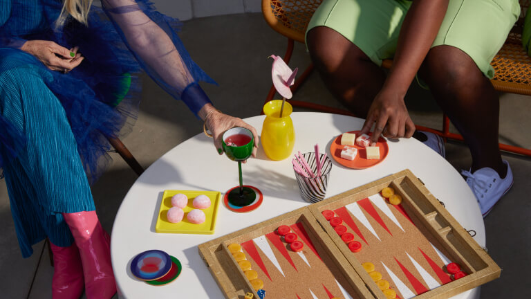 A Black woman and White woman share snacks and drinks over a board game.