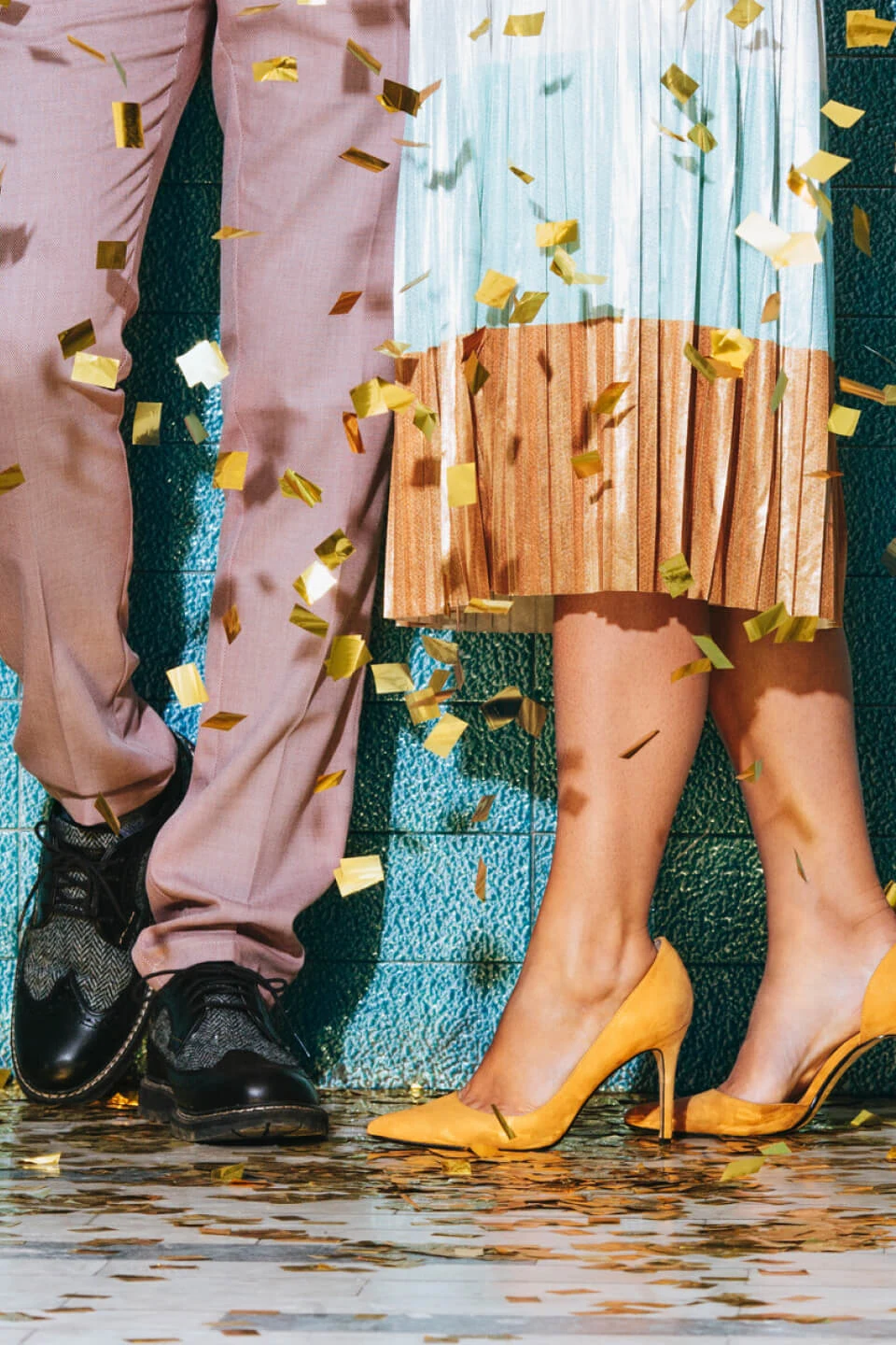 Pernas de duas pessoas próximas uma da outra contra uma parede azul, cercadas por confetes. A pessoa à esquerda veste uma calça na cor salmão e sapato social preto. A pessoa da direita usa uma saia bicolor e sapato de salto amarelo.
