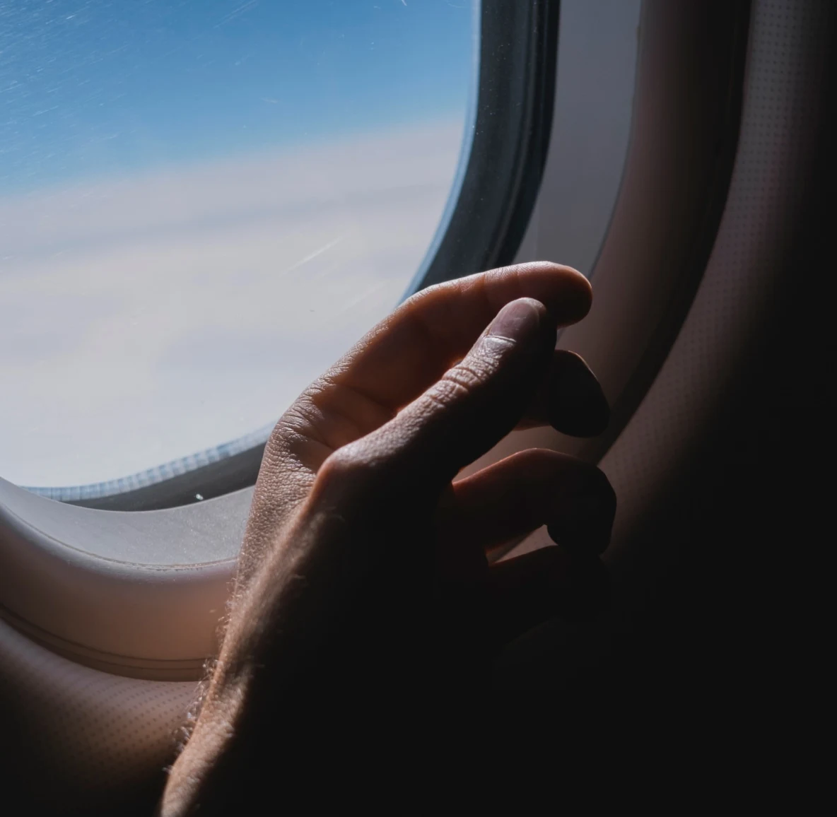 A hand in the shadow of an airplane window, clouds and blue sky in the background
