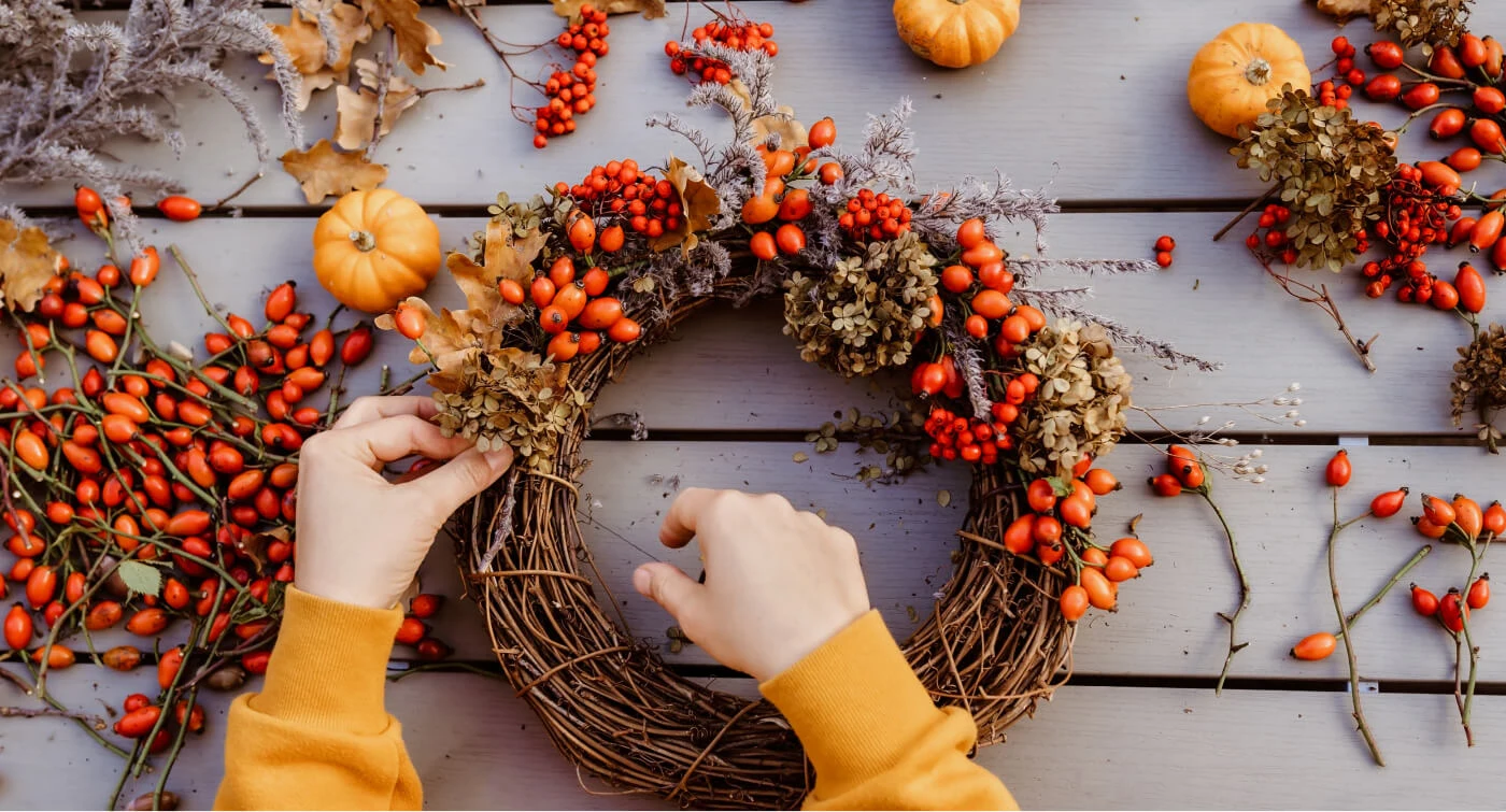 グレーの木製テーブルの上で、秋をテーマにしたリースを作っている両手を写した画像。リースは蔓で編まれた土台に、秋を思わせる枯葉、ドライフラワー、木の実で飾られている。テーブルにはローズヒップやミニカボチャ、その他の草花も置かれている。
