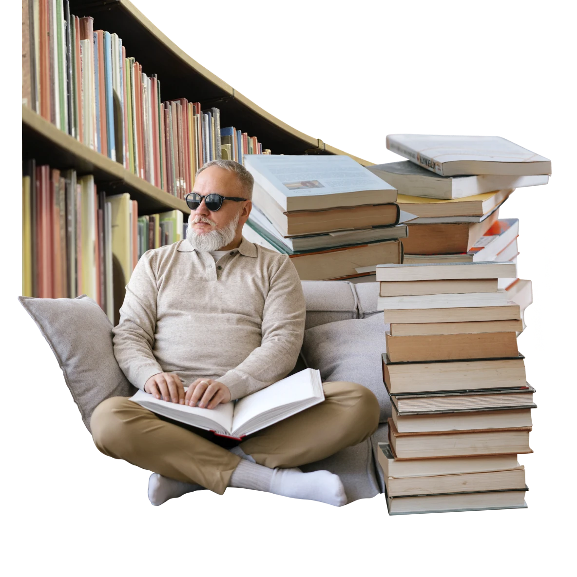 A visually impaired white man wearing sunglasses, sits cross-legged reading braille. He’s surrounded by stacks of books with a library bookshelf in the background.