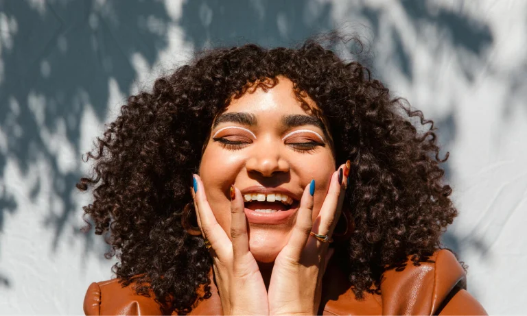 Black woman with curly hair laughs with her eyes closed and hands up to her cheeks