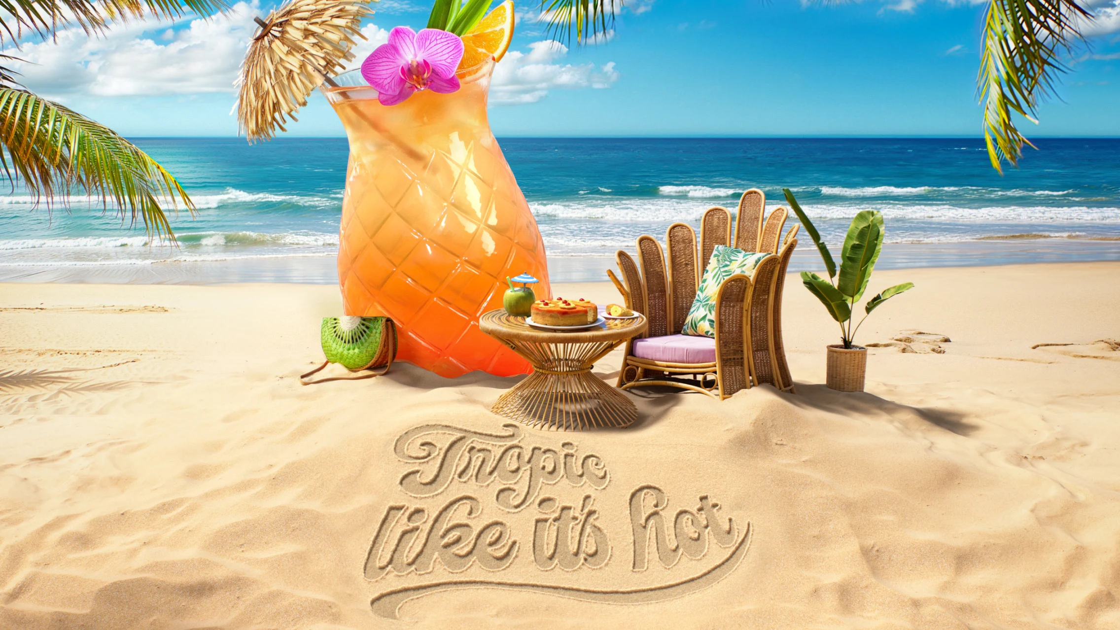 Life-sized tropical cocktail sits atop sandy shore in front of a bright blue ocean next to a rattan table and chair and green plants. “Tropic Like It’s Hot” is written in the sand in cursive font.