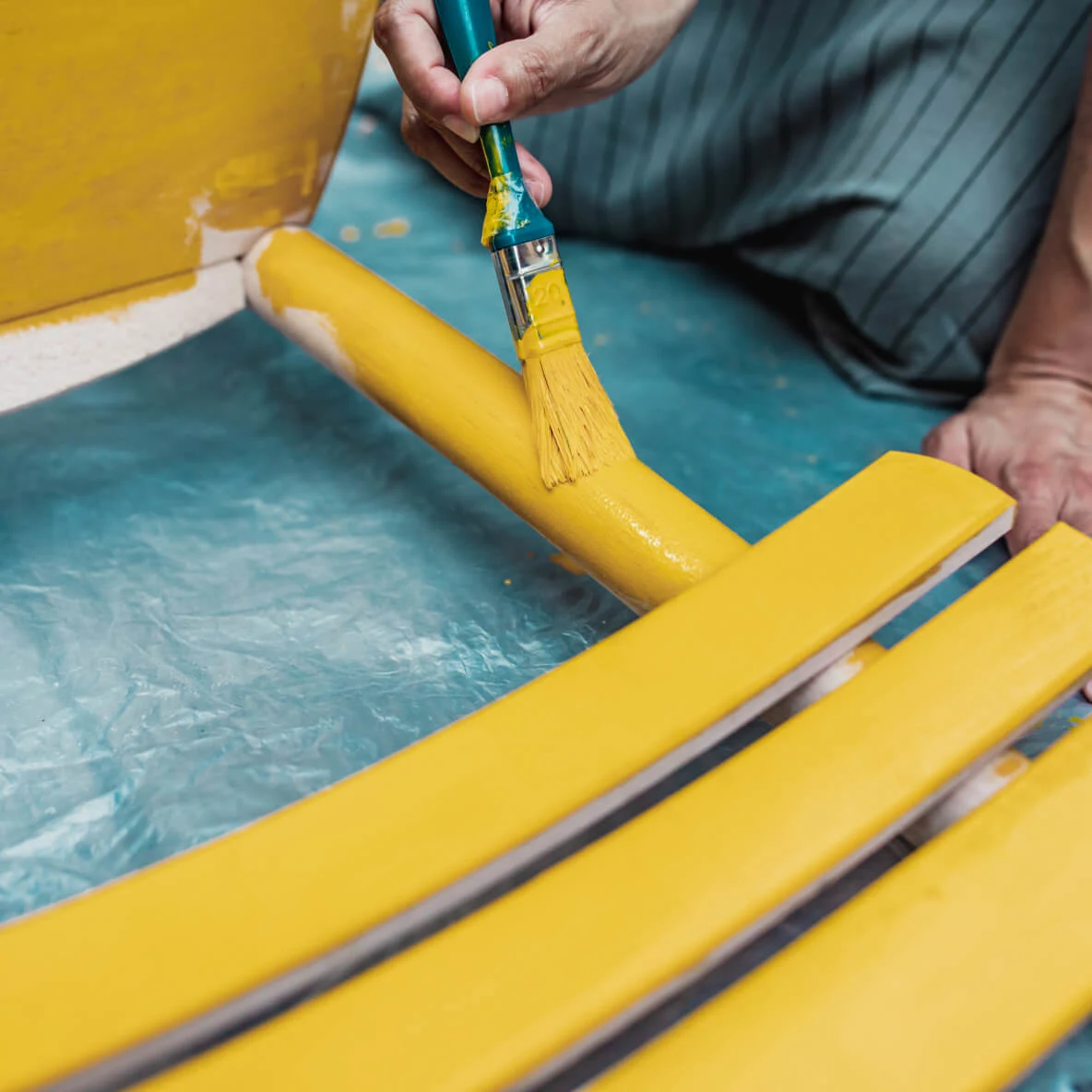 A pair of hands paints a wooden chair yellow with a brush. The chair is placed on a tarp to protect the ground. The focus is on the upcycling process rather than the person.