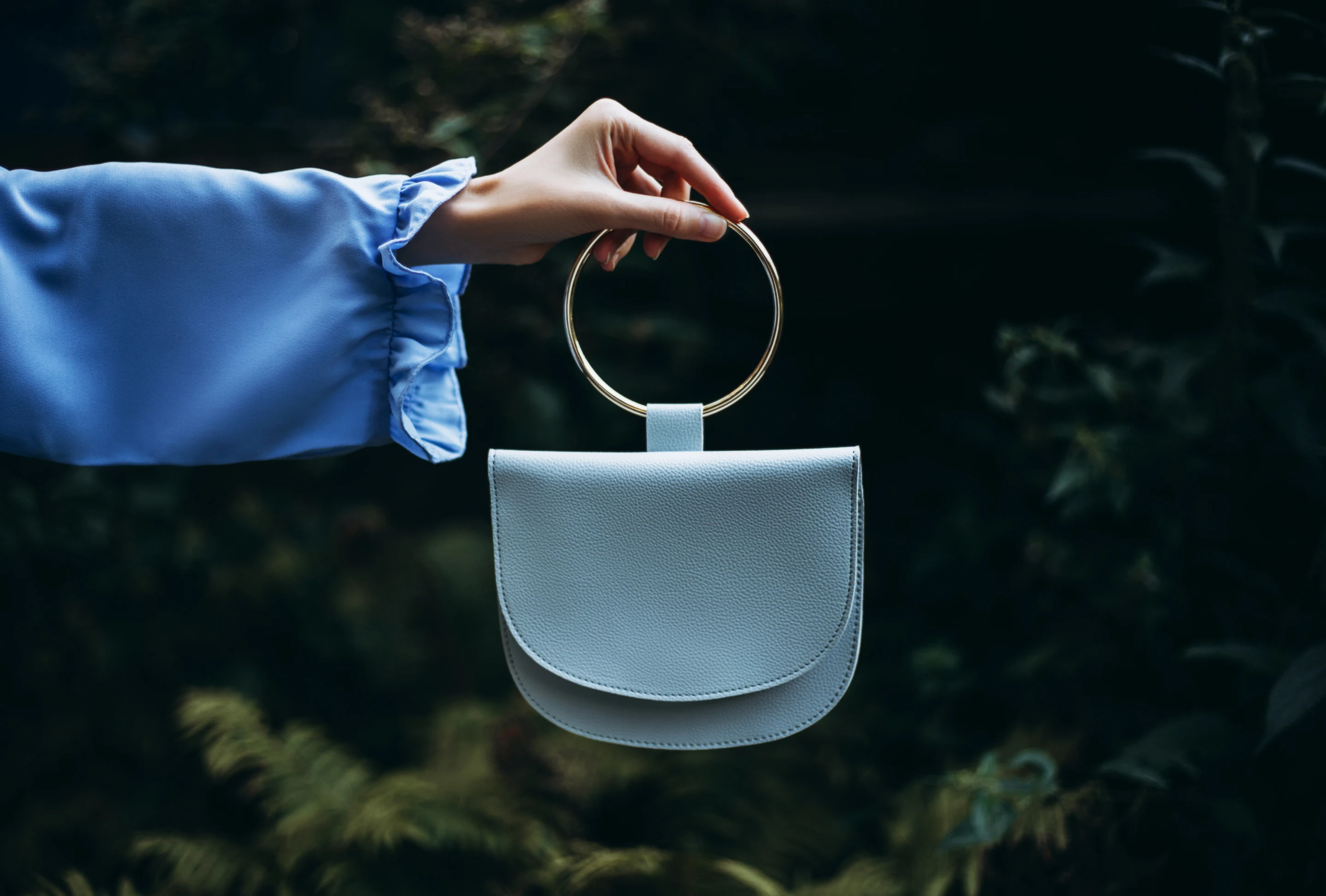 Une main dans une pochette bleue ample tient l��’anneau métallique argenté d’un petit sac à main bleu, avec une forêt floue en arrière-plan.