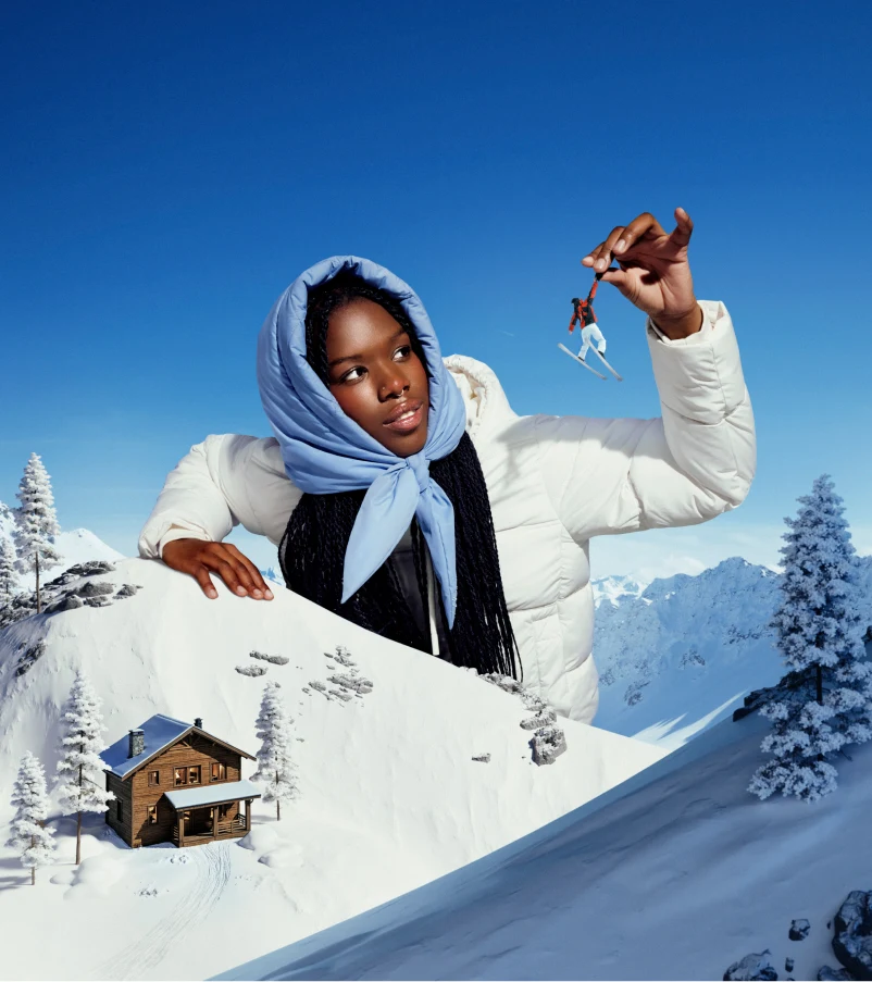 A woman stands against snowy rocks in a white puffy coat and periwinkle scarf. She is examining a tiny toy skier that she has picked up from a miniature scene of a ski resort.