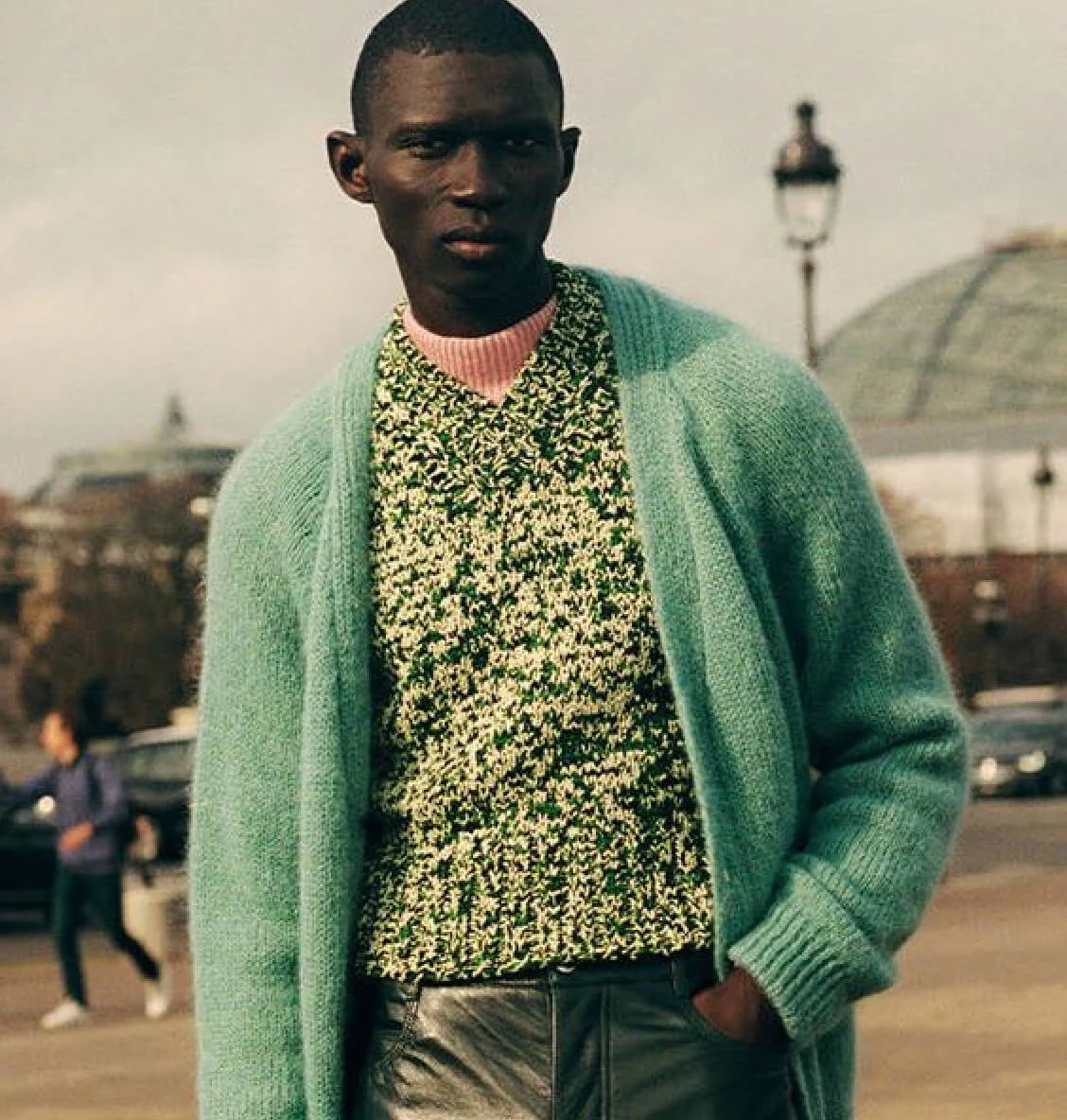 Black man stands on the street wearing leather pants and green cardigan over a green sweater and pink turtleneck. A lamppost, cars and pedestrian are visible in the background.