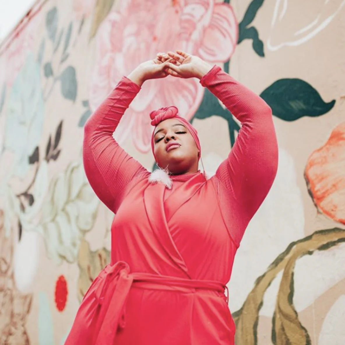 Woman in salmon colored robe tied in the front and matching turban with her hands clasped above her head in front of a  wall with a floral mural