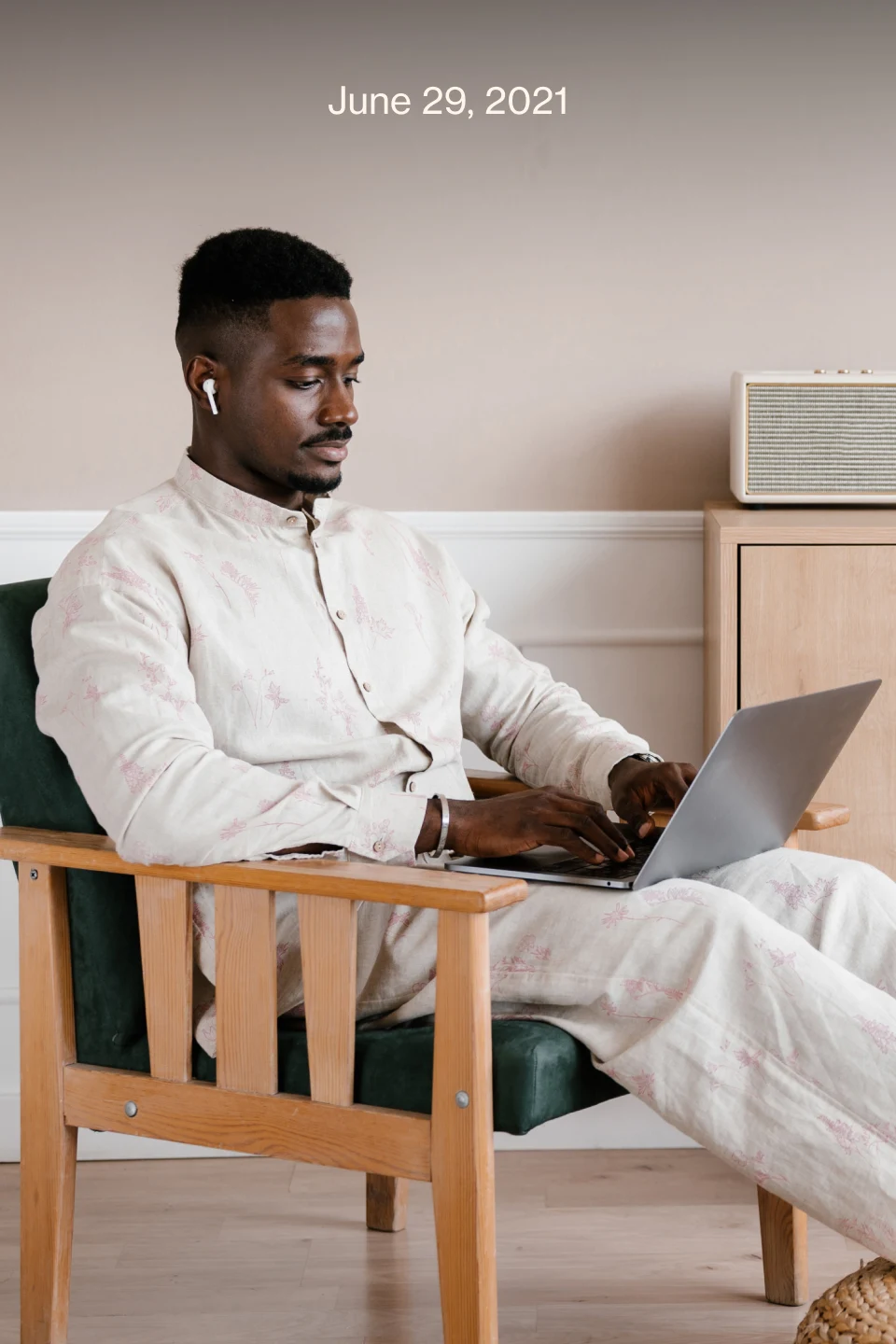 Black man wearing a beige shirt and pants with earphones sitting in a chair typing on a computer.