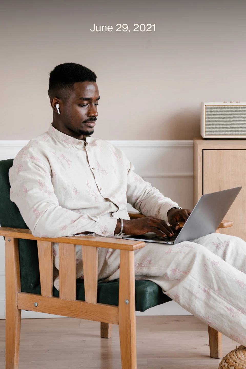 Black man wearing a beige shirt and trousers, with earphones, sitting in a chair, typing on a computer.