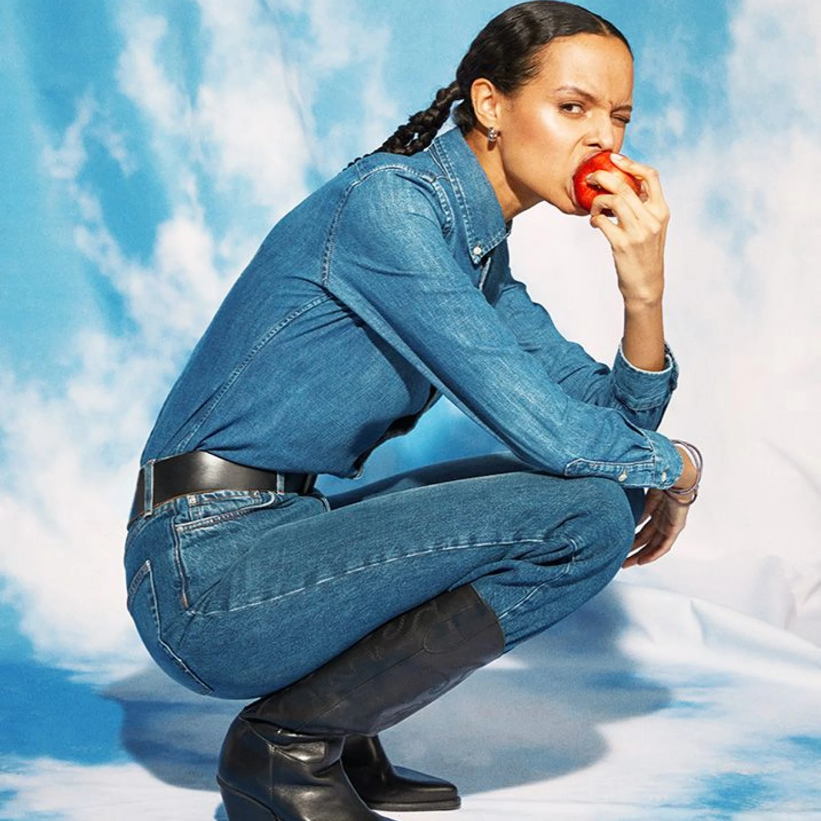 A women dressed in head to toe denim crouches down whilst biting into an apple 