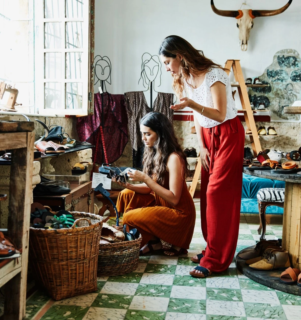 Duas mulheres com cabelo comprido e escuro analisam um par de sandálias numa loja de roupa