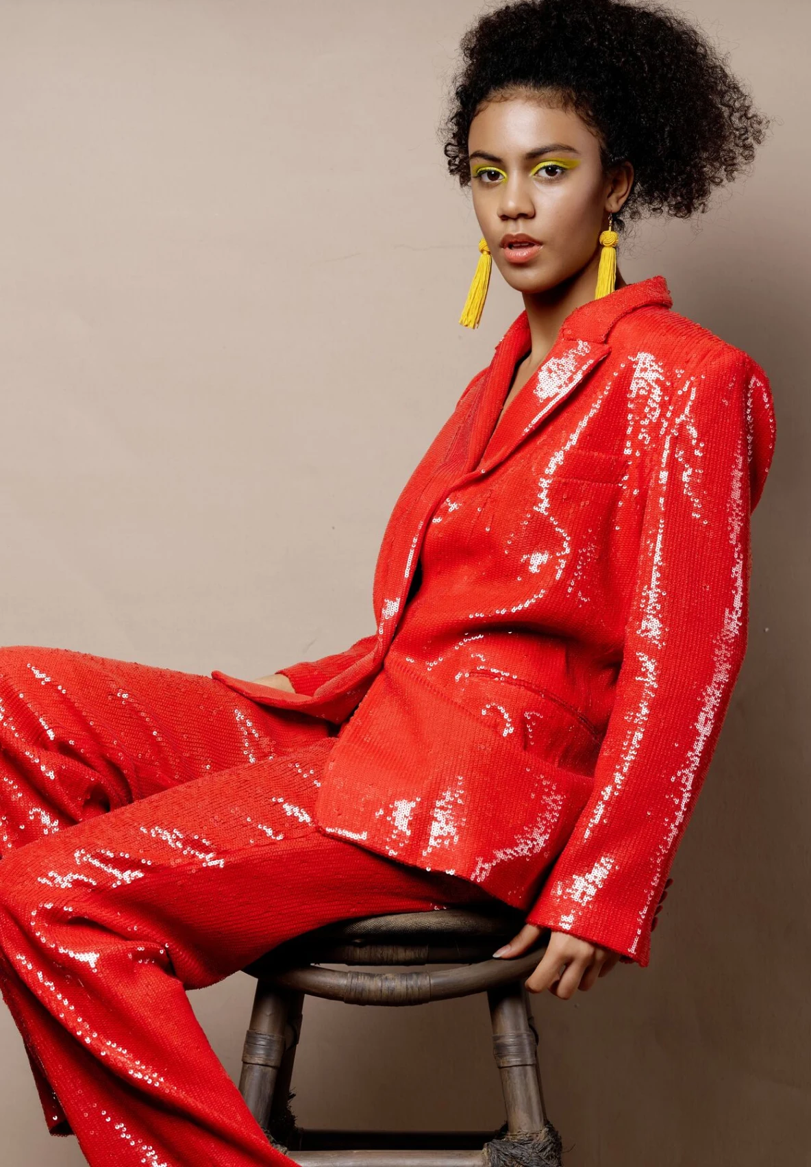Image of a woman sitting on a wooden stool, wearing a red sequined suit. She is also wearing bright yellow dangling earrings and bright yellow liquid eyeshadow, with her hair tied up in a ponytail. 