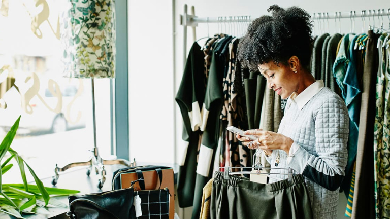 A Black woman with coily hair checking her mobile phone while shopping in a small clothing boutique.