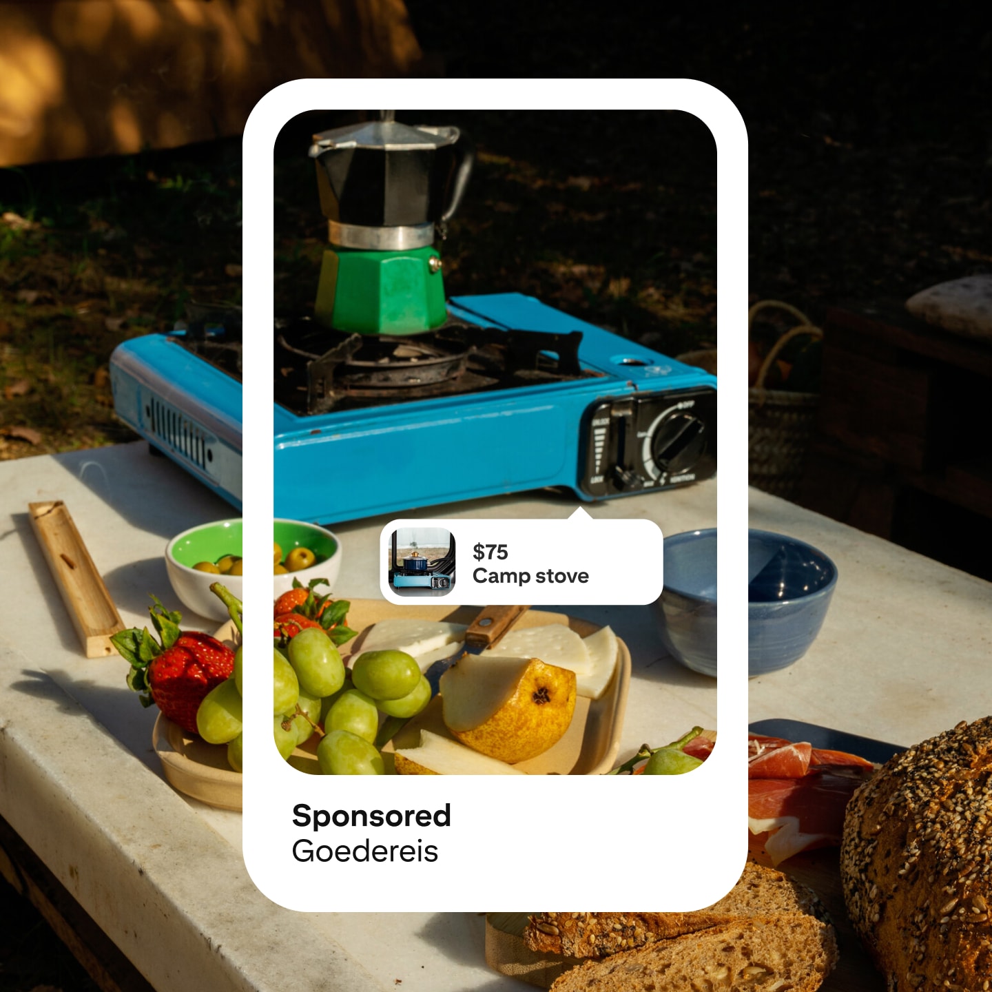 An image of a Pinterest ad mockup displaying a blue and silver portable camp stove priced at $75, labeled "Camp stove" and sponsored by "Goedereis." Various fruits and food items are spread around the stove in an outdoor setting.