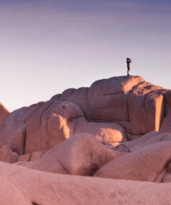 Una persona in lontananza che guarda il tramonto dall'alto di una grande formazione rocciosa nel deserto. 